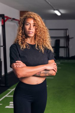 Uzun kıvırcık saçlı, kendine güvenen sporcu kadın spor salonunda kollarını kavuşturup kameraya, fitness konseptine ve aktif yaşam tarzına bakıyor.