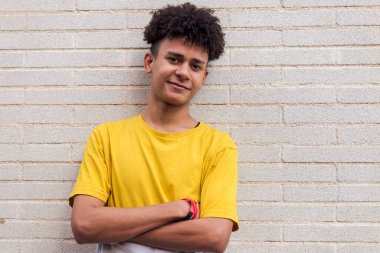 Neşeli Afro-Amerikan erkek, günlük kıyafetlerle mutlu bir şekilde gülümsüyor. Tuğla duvara yaslanmış kollarıyla kameraya, gençlik ve kentsel yaşam tarzına bakıyor. Metinler için kopya alanı.