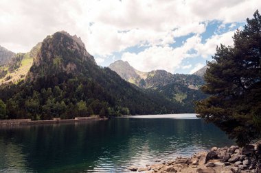 Büyük yeşil dağlar ve kozalaklı ağaçlarla çevrili sakin göl manzarası Aiguestortes I Estany of Saint Maurici Ulusal Parkı 'nda bulutlu gökyüzüne karşı
