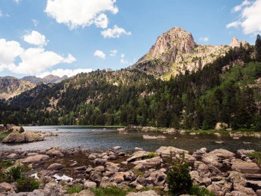Önünde bir göl olan dağ sırası göl kayalar ve ağaçlarla çevrilidir, gökyüzü açık ve mavidir.