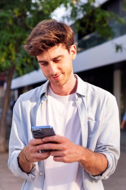 Gülümseyen adam sokakta yürürken cep telefonunu kullanıyor, beyaz bir gömlek giyiyor ve telefonuna odaklanmış durumda, iletişim teknolojisi ve kentsel yaşam tarzı, metin için kopyalama alanı