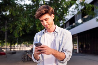 Gülümseyen adam sokakta yürürken cep telefonunu kullanıyor, beyaz bir gömlek giyiyor ve telefonuna odaklanmış durumda, iletişim teknolojisi ve kentsel yaşam tarzı, metin için kopyalama alanı