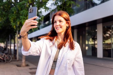 Gülümseyen genç bir kadın cep telefonuyla selfie çekiyor, mutlu ve anın tadını çıkarıyor, iletişim teknolojisi ve şehir yaşam tarzı kavramı.
