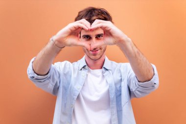 Turuncu duvarın önünde duran genç adam kameraya bakarken kalp şeklinde bir kalp yapıyor, mavi gömlek ve beyaz tişört giyiyor, aşk kavramı, metin için metin alanı