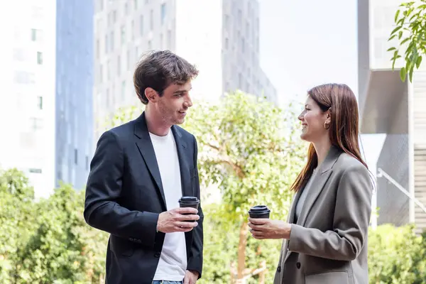 Gülümseyen bir adam ve bir kadın işyeri binalarının yakınındaki bir parkta kahve fincanı alırken konuşuyor, gülümsüyor ve şirketle, iş ve kentsel yaşam tarzıyla eğleniyorlar.