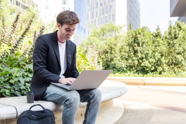 Ofis binalarının yakınındaki bir parkın bankında dizüstü bilgisayar kullanan gülümseyen genç adam, uzaktan çalışma için iş ve teknoloji kavramı