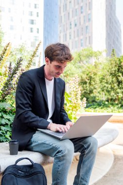 Ofis binalarının yakınındaki bir parkın bankında dizüstü bilgisayar kullanan gülümseyen genç adam, uzaktan çalışma için iş ve teknoloji kavramı