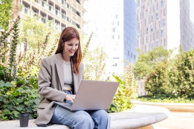 Ofis binalarının yakınındaki bir parkın bankında dizüstü bilgisayar kullanan gülümseyen genç bir kadın, uzaktan çalışma için iş ve teknoloji kavramı