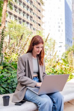 Ofis binalarının yakınındaki bir parkın bankında dizüstü bilgisayar kullanan gülümseyen genç bir kadın, uzaktan çalışma için iş ve teknoloji kavramı