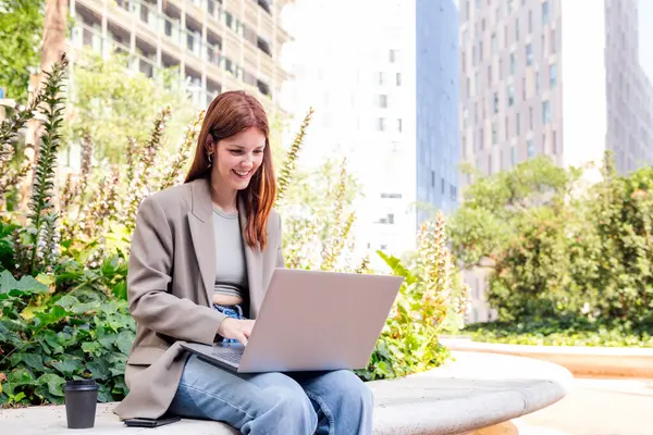 Ofis binalarının yakınındaki bir parkın bankında dizüstü bilgisayar kullanan gülümseyen genç bir kadın, uzaktan çalışma için iş ve teknoloji kavramı