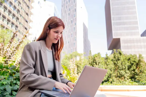 Ofis binalarının yakınındaki bir parkın bankında dizüstü bilgisayar kullanan gülümseyen genç bir kadın, uzaktan çalışma için iş ve teknoloji kavramı