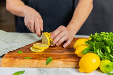 Tahta kesme tahtasındaki taze limonları dilimleyen ellerin yakın çekimi. İçinde bir deste limon, nane yaprağı ve mermer yüzeyde yeşil bir havlu var..