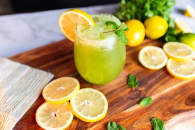 Buzlu, naneli ve limonlu bir bardakta servis edilen ferahlatıcı yeşil limonata, arka planda taze limonlar ve otlarla dolu ahşap bir tahtanın üzerine yerleştirilmiş..