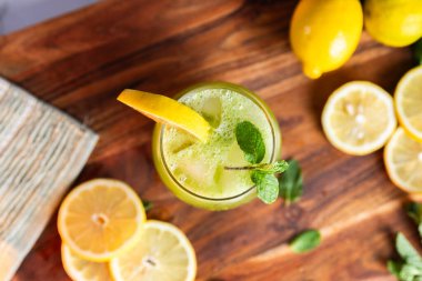 Buzlu, naneli ve limonlu bir bardakta servis edilen ferahlatıcı yeşil limonata, arka planda taze limonlar ve otlarla dolu ahşap bir tahtanın üzerine yerleştirilmiş..