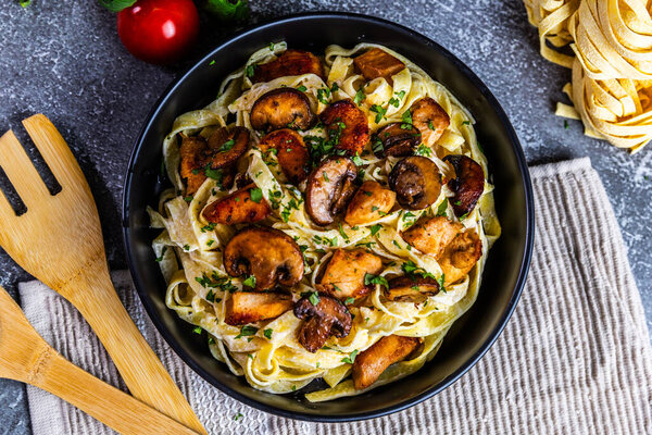 A delicious serving of creamy tagliatelle pasta topped with golden chicken and sauted mushrooms, garnished with fresh parsley. Presented in a black bowl on a rustic background.