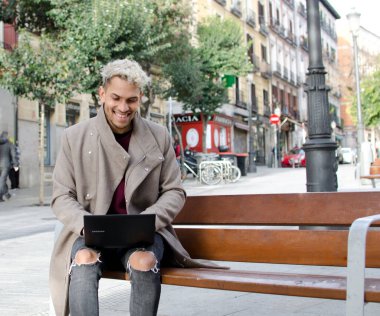 Yakın plan genç, mutlu ve zarif bir adam sokakta dizüstü bilgisayarla çalışıyor.