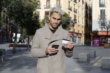 Yakın plan genç, mutlu ve zarif bir adam, sokakta yürüyor, cep telefonu ve kredi kartıyla online alışveriş yapıyor..