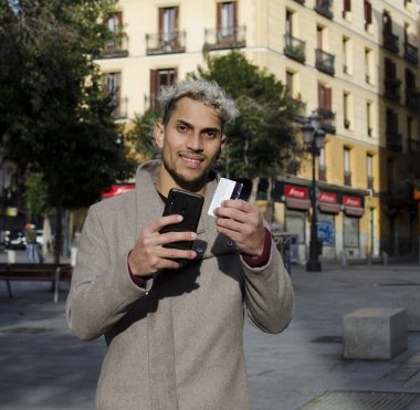 Yakın plan genç, mutlu ve zarif bir adam, sokakta yürüyor, cep telefonu ve kredi kartıyla online alışveriş yapıyor..