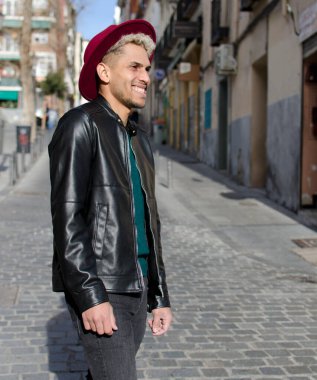 Deri ceketli, kırmızı şapkalı, sokakta gülümseyen genç bir manken.