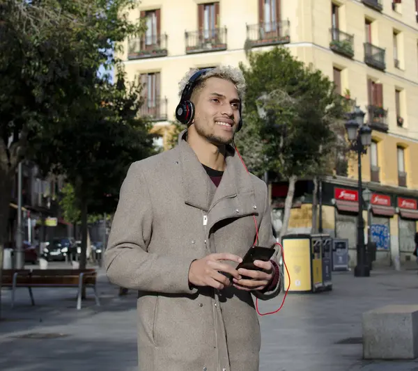Kulaklıklı genç adam akıllı telefonu elinde tutuyor ve şehirde müzik dinliyor.
