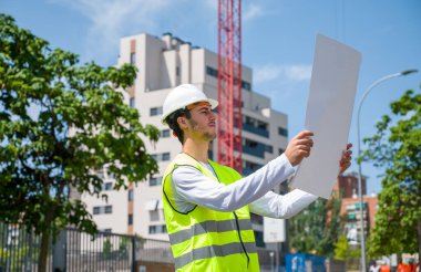 Güvenlik kaskı ve yansıtıcı yeleği olan profesyonel erkek inşaat işçisi. İnşaat alanındaki çalışma planını kontrol ediyor.