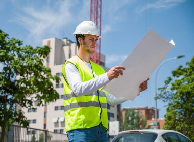 Güvenlik kaskı ve yansıtıcı yeleği olan profesyonel erkek inşaat işçisi. İnşaat alanındaki çalışma planını kontrol ediyor.