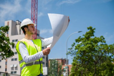 Güvenlik kaskı ve yansıtıcı yeleği olan profesyonel erkek inşaat işçisi. İnşaat alanındaki çalışma planını kontrol ediyor.