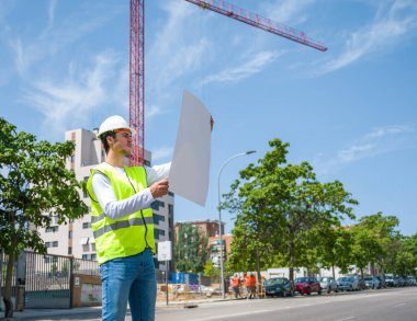 Güvenlik kaskı ve yansıtıcı yeleği olan profesyonel erkek inşaat işçisi. İnşaat alanındaki çalışma planını kontrol ediyor.