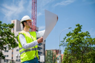 Güvenlik kaskı ve yansıtıcı yeleği olan profesyonel erkek inşaat işçisi. İnşaat alanındaki çalışma planını kontrol ediyor.