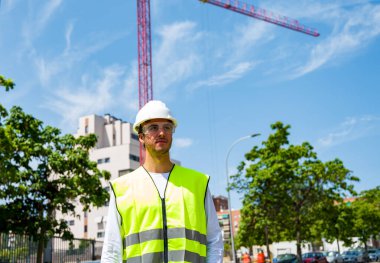 Güvenlik kaskı ve yansıtıcı yeleği olan profesyonel erkek inşaat işçisi. İnşaat alanında arka planda inşaat çalışması var.