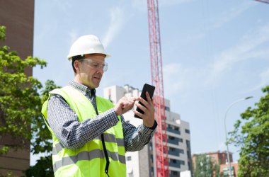 Güvenlik kaskı, yansıtıcı yelek ve güvenlik gözlükleriyle inşaat alanında tablet kullanan profesyonel bir inşaat işçisi.