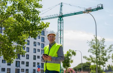 Güvenlik kaskı, yansıtıcı yelek ve güvenlik gözlükleriyle inşaat alanında tablet kullanan profesyonel bir inşaat işçisi.