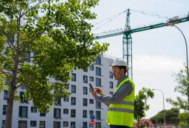 Güvenlik kaskı, yansıtıcı yelek ve güvenlik gözlükleriyle inşaat alanında tablet kullanan profesyonel bir inşaat işçisi.