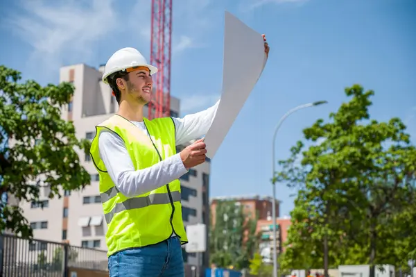 Güvenlik kaskı ve yansıtıcı yeleği olan profesyonel erkek inşaat işçisi. İnşaat alanındaki çalışma planını kontrol ediyor.