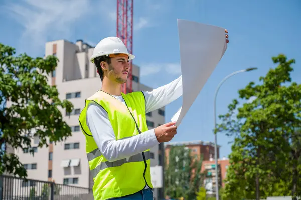 Güvenlik kaskı ve yansıtıcı yeleği olan profesyonel erkek inşaat işçisi. İnşaat alanındaki çalışma planını kontrol ediyor.