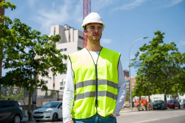 Güvenlik kaskı ve yansıtıcı yeleği olan profesyonel erkek inşaat işçisi. İnşaat alanında arka planda inşaat çalışması var.