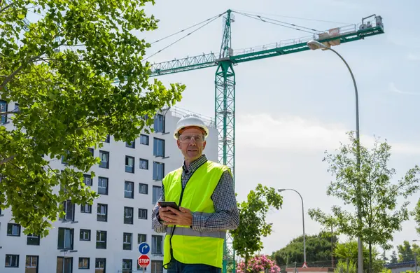 Güvenlik kaskı, yansıtıcı yelek ve güvenlik gözlükleriyle inşaat alanında tablet kullanan profesyonel bir inşaat işçisi.