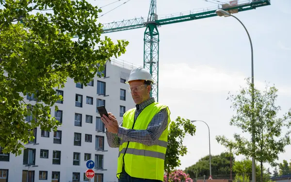 Güvenlik kaskı, yansıtıcı yelek ve güvenlik gözlükleriyle inşaat alanında tablet kullanan profesyonel bir inşaat işçisi.