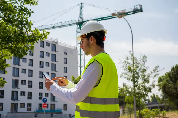 Güvenlik kaskı ve yansıtıcı yeleği olan profesyonel erkek inşaat işçisi. İnşaat alanında arka planda inşaat çalışması var.