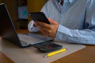 Ofiste akıllı telefon ve laptop kullanan bir iş adamı. Adam sosyal medyayı tarıyor, e-postaları kontrol ediyor, iş işlemlerini yönetiyor, uzaktan çalışıyor. Modern teknoloji işi kolaylaştırır. Etkin zaman yönetimi.