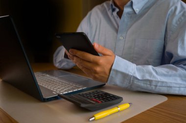 Ofiste akıllı telefon ve laptop kullanan bir iş adamı. Adam sosyal medyayı tarıyor, e-postaları kontrol ediyor, iş işlemlerini yönetiyor, uzaktan çalışıyor. Modern teknoloji işi kolaylaştırır. Etkin zaman yönetimi.