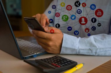 Ofiste akıllı telefon ve laptop kullanan bir iş adamı. Adam sosyal medyayı tarıyor, e-postaları kontrol ediyor, iş işlemlerini yönetiyor, uzaktan çalışıyor. Modern teknoloji işi kolaylaştırır. Etkin zaman yönetimi.