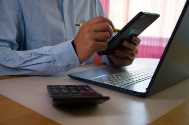 Ofiste akıllı telefon ve laptop kullanan bir iş adamı. Adam sosyal medyayı tarıyor, e-postaları kontrol ediyor, iş işlemlerini yönetiyor, uzaktan çalışıyor. Modern teknoloji işi kolaylaştırır. Etkin zaman yönetimi.