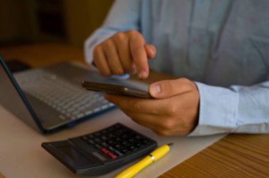 Ofiste akıllı telefon ve laptop kullanan bir iş adamı. Adam sosyal medyayı tarıyor, e-postaları kontrol ediyor, iş işlemlerini yönetiyor, uzaktan çalışıyor. Modern teknoloji işi kolaylaştırır. Etkin zaman yönetimi.