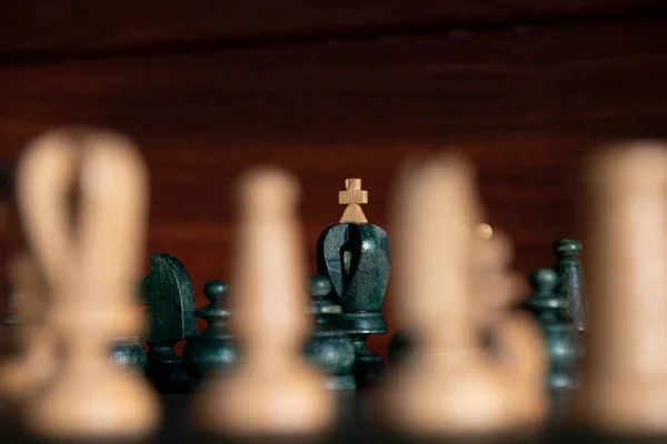 Black king with white cross in background between white chess pieces out of focus with brown wooden background. Concept: chess