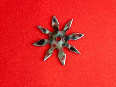 Kırmızı arka planda askeri renklerle boyanmış yıldız şeklinde bir shuriken. Shuriken 'daki yazıda Çin yapımı olduğu yazıyor..