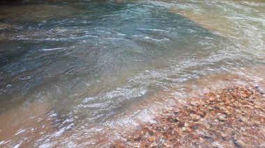 Nehirdeki dalgalar pek çok taşın arasından akar. Renkli yaz mevsiminden parlak güneş ışığına. Arka plandaki parlak çakıl taşı, su yüzeyinin altındaki çakıl taşlarının soyut bir şekliyle derenin dibi..