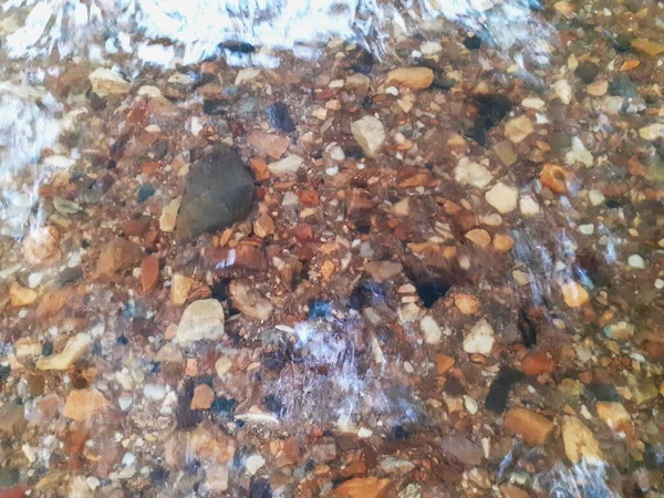 Ripples in the river flow through many stones, colorful summer to bright sunlight. The background shiny pebble, with A bottom of stream In an abstract, form Of pebbles below water surface.
