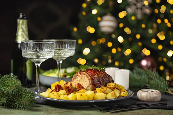 Christmas or New Year dinner table. Baked duck roulade stuffed with meat and oranges, baked potato, salad, champagne coupe glasses in front of Christmas tree and burning candles.