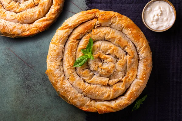 stock image Top view of Rolled Burek - savoury round pie fith meat feeling, decorated with sesam seedes, yogurt. Dark table table.sna